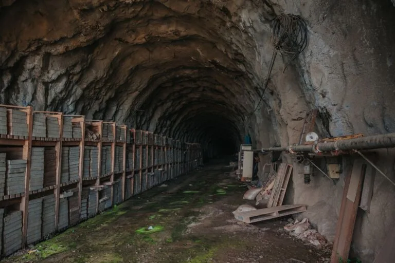 Galleria mineraria con pareti rocciose scure, cassoni metallici arrugginiti e tubi industriali su pavimento muschiato.