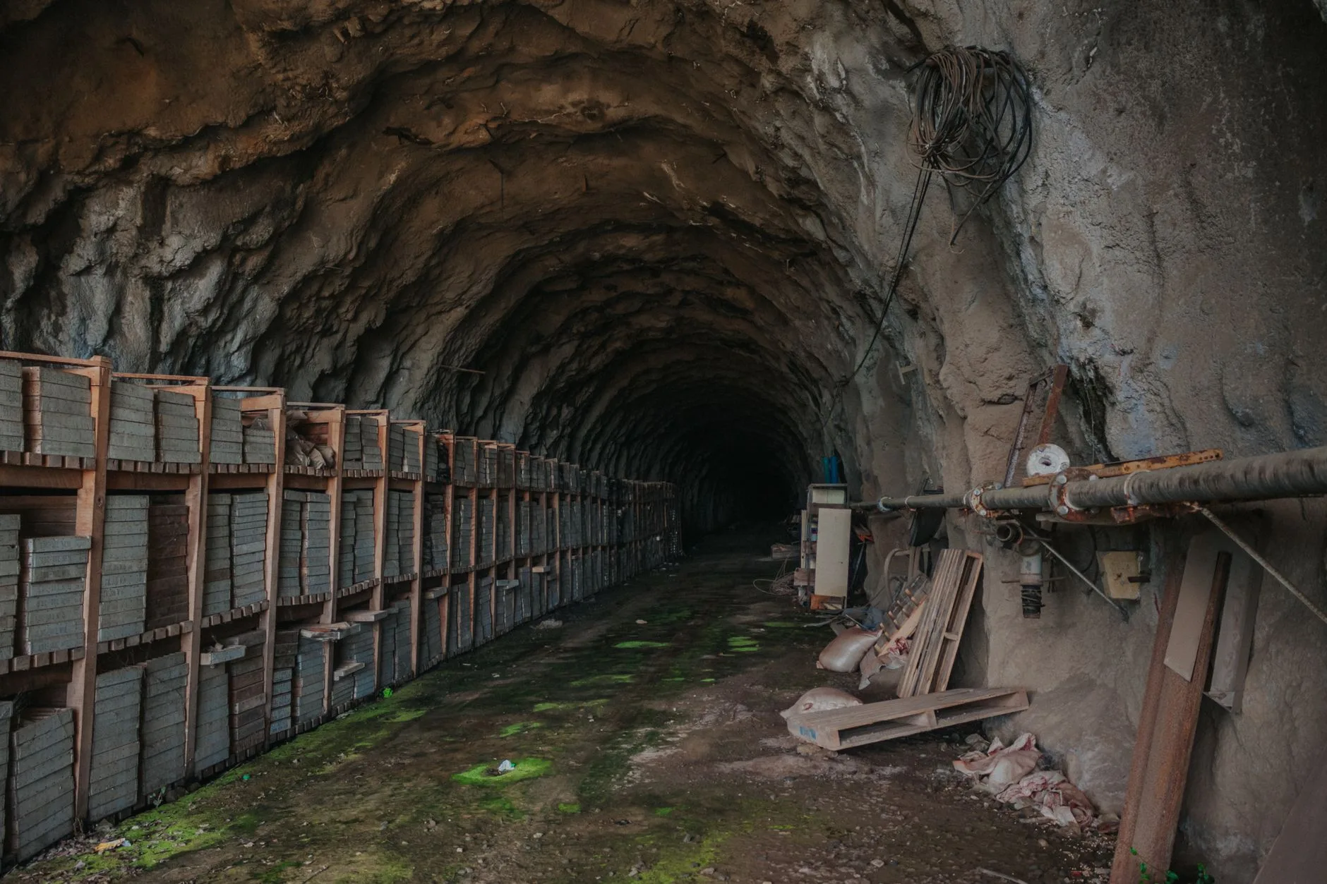 Galleria mineraria con pareti rocciose scure, cassoni metallici arrugginiti e tubi industriali su pavimento muschiato.