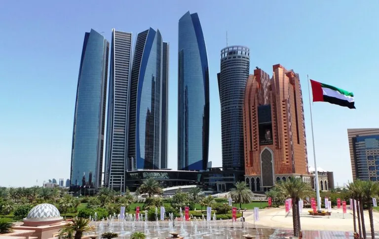 Grattacieli moderni con bandiera emiratina, piazza con fontana e palme in un centro urbano del Golfo.