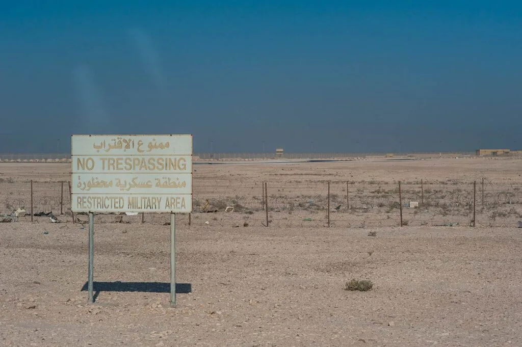 Cartello bilingue che vieta l'accesso in area militare ristretta nel deserto, con recinzione e territorio arido.