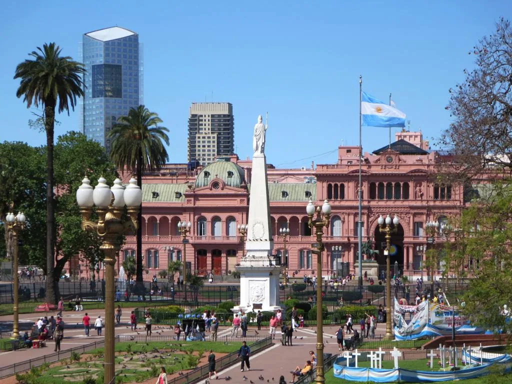 Piazza principale di Buenos Aires con palazzo presidenziale rosa, monumento centrale, palme e bandiera argentina al vento.