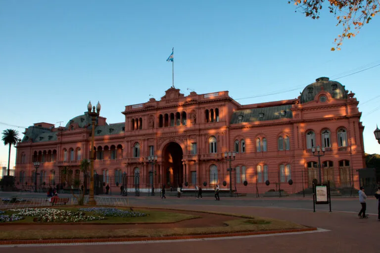 Palazzo presidenziale rosa illuminato al tramonto con bandiera argentina in cima, circondato da una piazza con persone.
