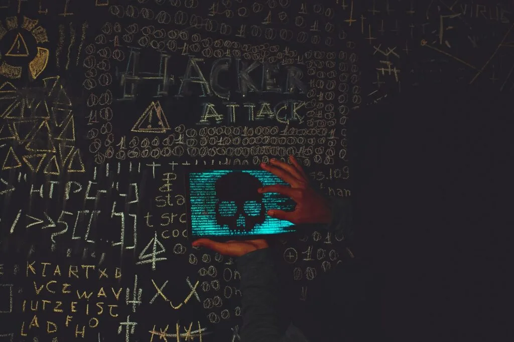 Mano su tablet che mostra un teschio blu circondato da simboli di codice e scritta 'Hacker Attack' su fondo scuro.