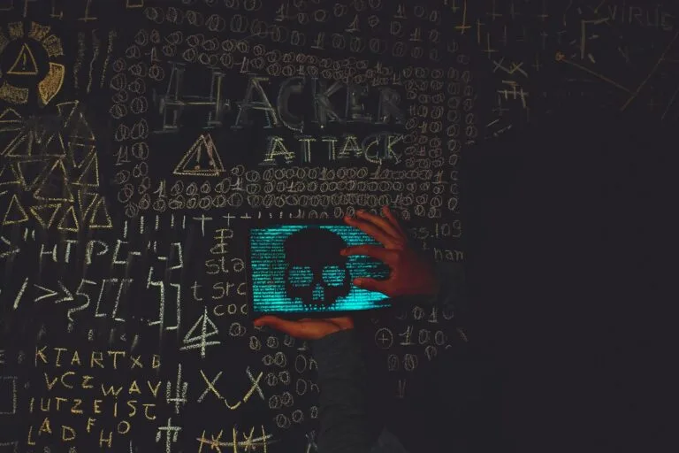 Mano su tablet che mostra un teschio blu circondato da simboli di codice e scritta 'Hacker Attack' su fondo scuro.