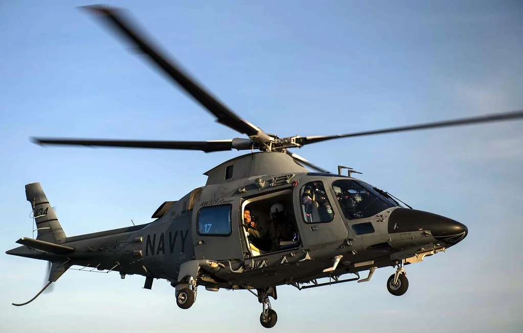 Elicottero militare in volo con livrea della marina, rotori in movimento contro cielo azzurro.