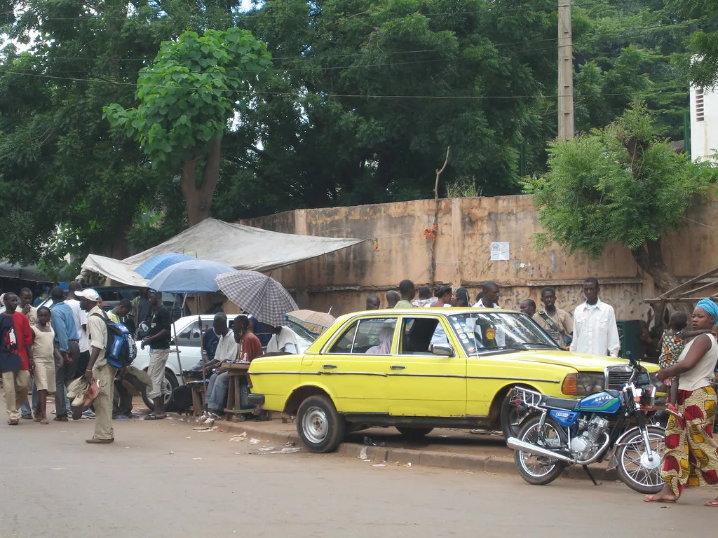 Taxi giallo Mercedes W123 circondato da persone in una strada di Bamako con tende e muri diroccati.