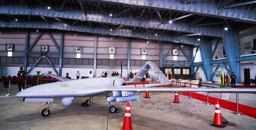 Drone tattico grigio esposto in un hangar industriale circondato da personale e barriere di sicurezza.