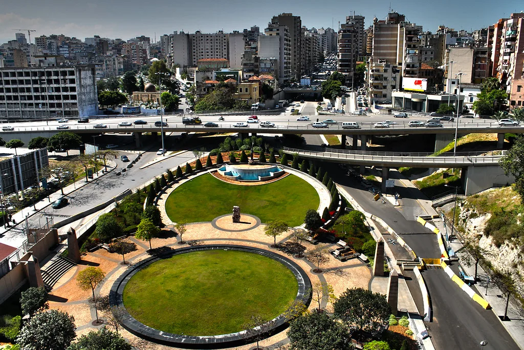 Parco urbano moderno con piscina e aree verdi circondate da edifici residenziali e infrastrutture stradali elevate.
