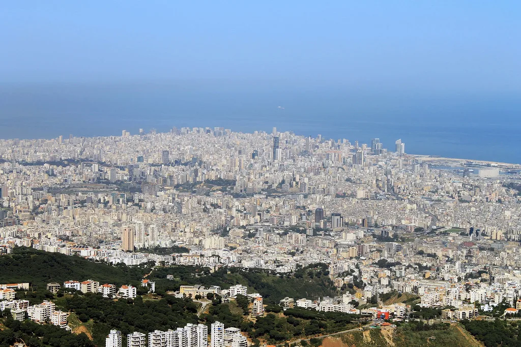 Città costiera mediorientale densamente costruita con quartieri residenziali in primo piano e mare blu sullo sfondo.