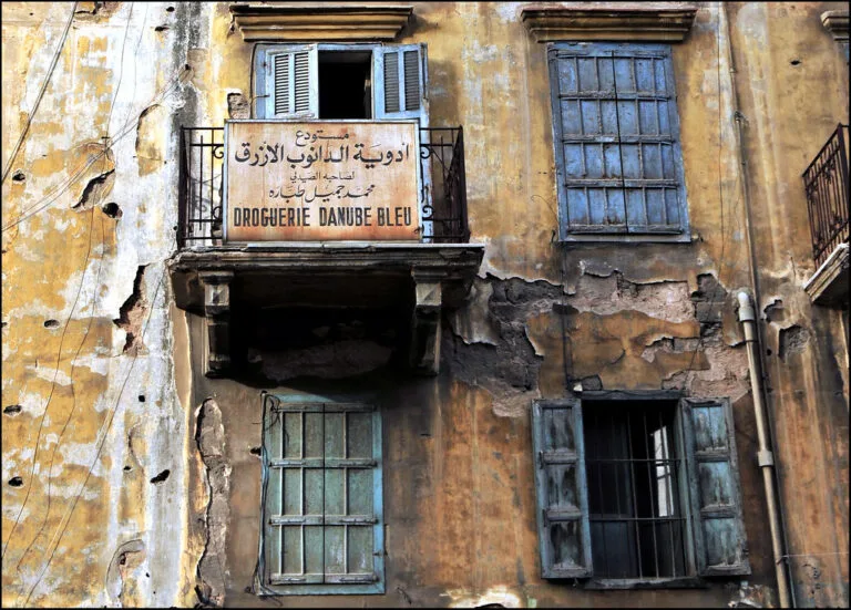 Edificio libanese deteriorato con insegna di farmacia in arabo e francese, balcone e finestre blu.