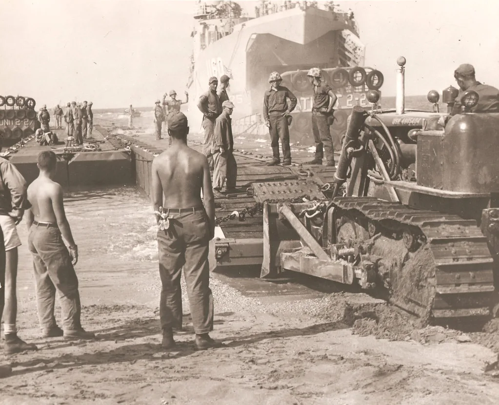 Soldati e operai lavorano attorno a un mezzo militare cingolato su una spiaggia, con una nave da guerra sullo sfondo.