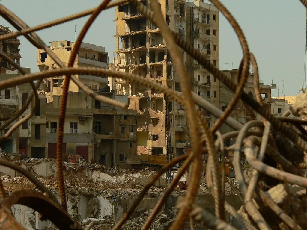 Edifici residenziali danneggiati da bombardamenti ripresi attraverso ferri arrugginiti contorti in primo piano.