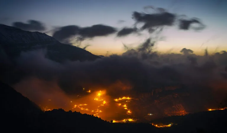 Città illuminata dalle fiamme nella notte con montagne scure in primo piano e nuvole grigie al tramonto.