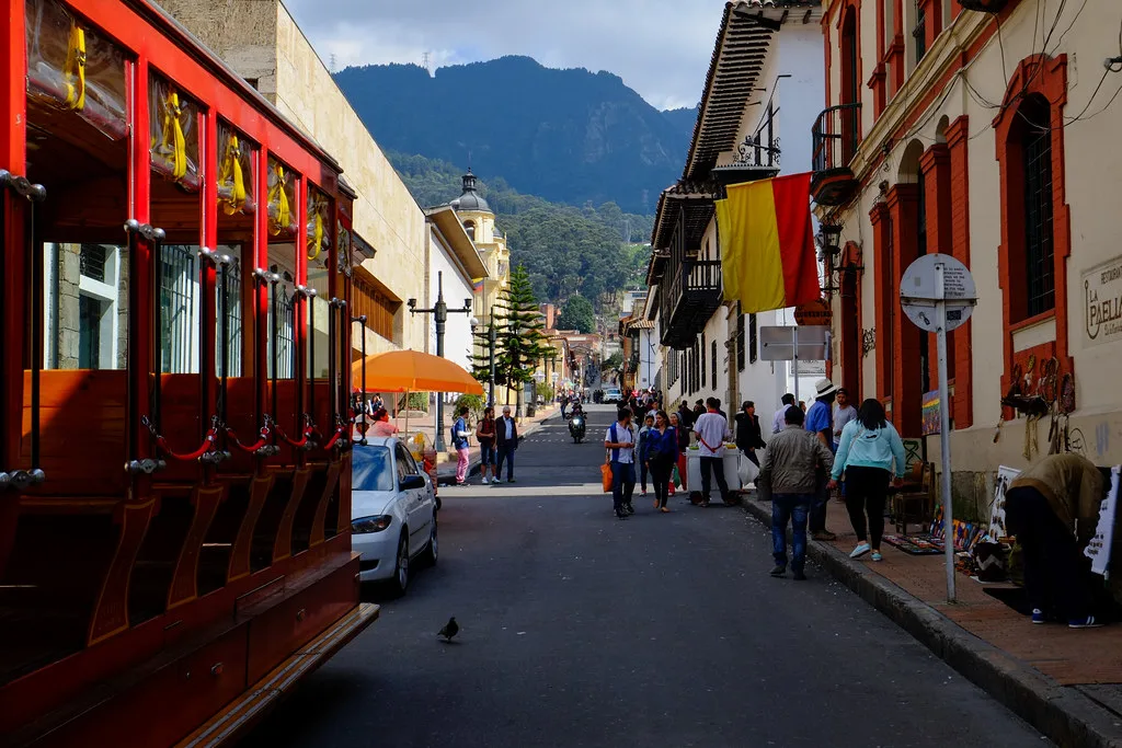 Strada coloniale del centro storico di Bogotá con edifici rossi affiancati, persone che passeggiando e montagne sullo sfondo.