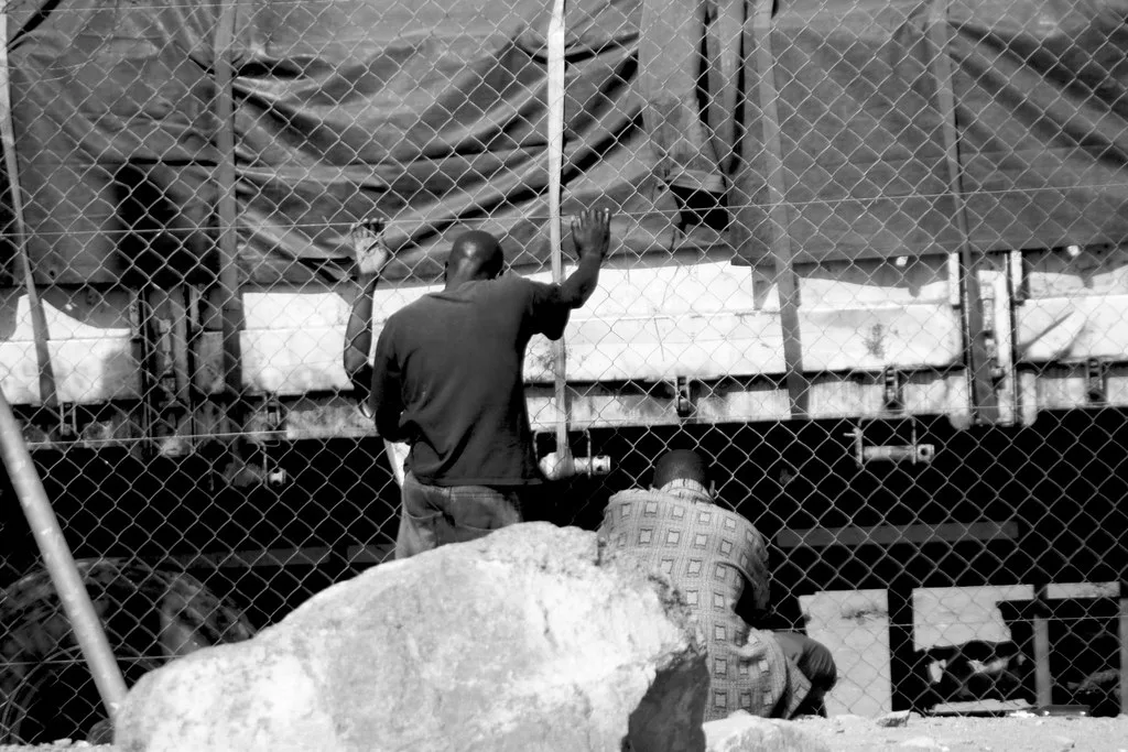 Uomo appoggia le mani su un recinto metallico in una zona povera con baracche sullo sfondo.