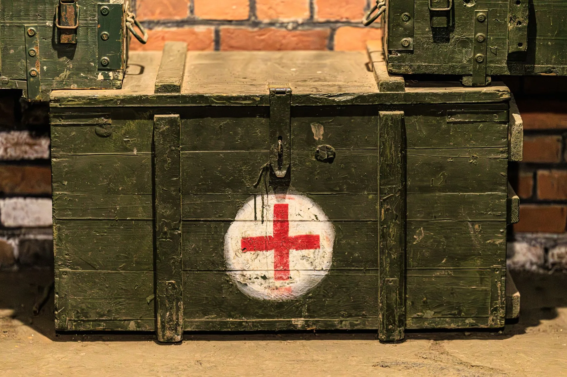 Cassone militare color verde oliva con croce rossa dipinta, elementi di fissaggio metallici e segni di usura.