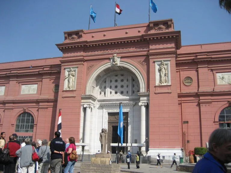 Edificio in terracotta con colonne bianche e portale ad arco, bandiere egiziana e internazionali, visitatori in piazza.