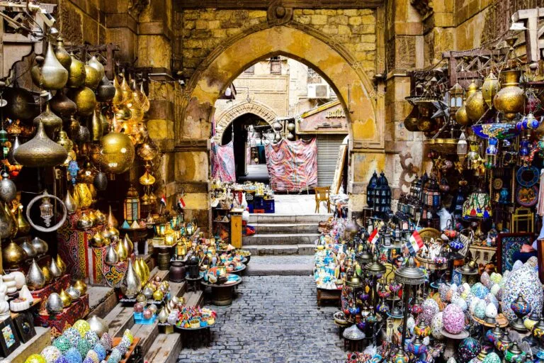 Bancarelle di artigianato in un mercato egiziano con lanterne dorate e ceramiche colorate sotto un arco di pietra.