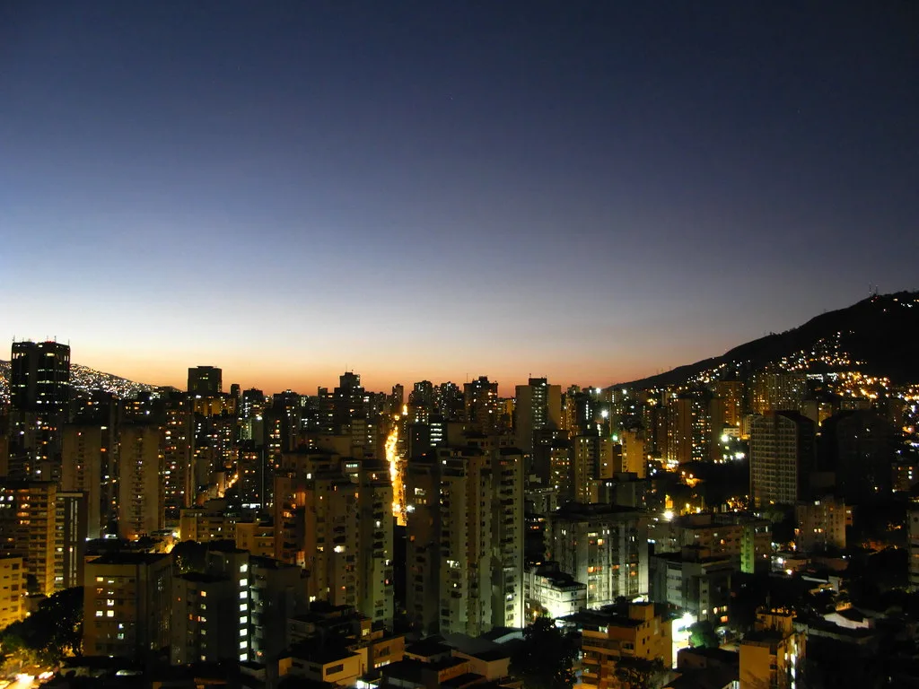 Skyline notturno di una città latinoamericana con grattacieli illuminati e montagna sullo sfondo al tramonto.