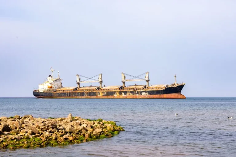 Nave cargo arrugginita in acque calme, vista dal lato con strutture di carico e scafo degradato color arancio.