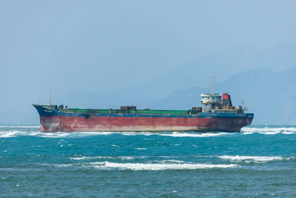 Nave cisterna dipinta di rosso e blu naviga in acque agitate con onde bianche e montagne sullo sfondo.