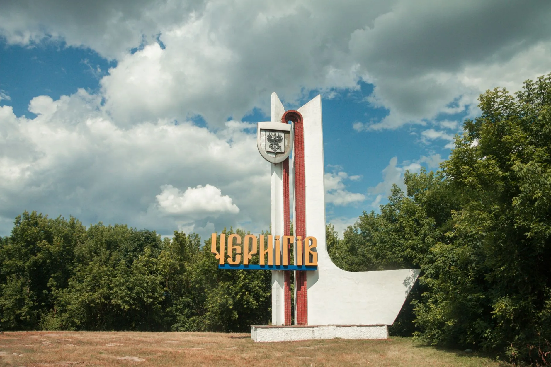 Monumento d'ingresso a Chernihiv con nome in oro su stele bianca a strisce rosse, circondato da alberi.