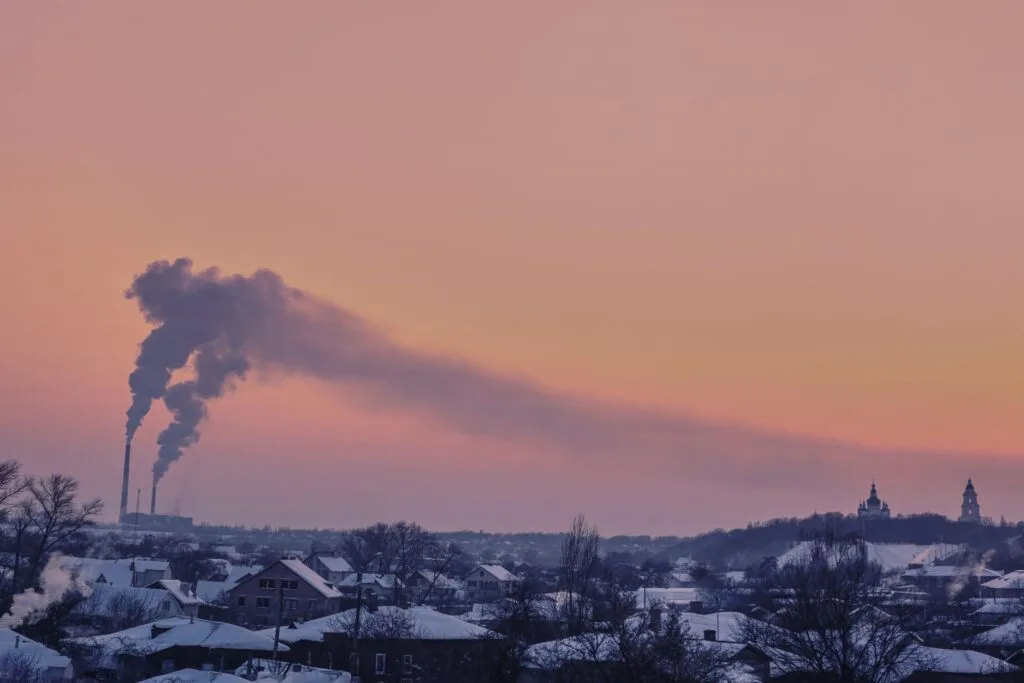 Camini industriali emettono denso fumo grigio su una città innevata al tramonto, con chiese sullo sfondo.