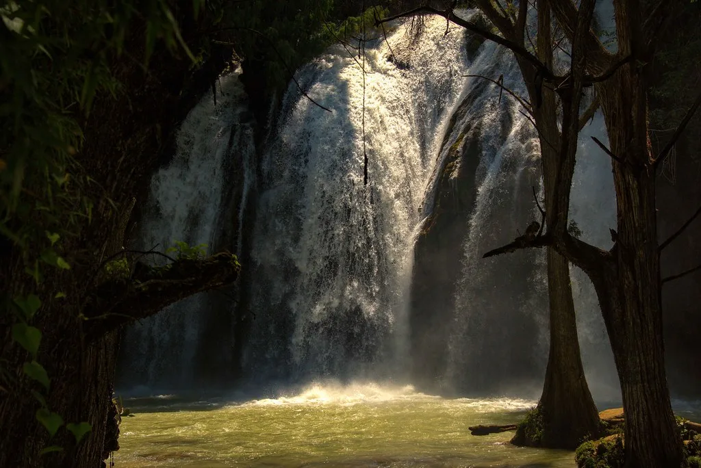 Cascata maestosa circondata da alberi tropicali con vasche d'acqua verde smeraldo alla base, in una zona di foresta pluviale.