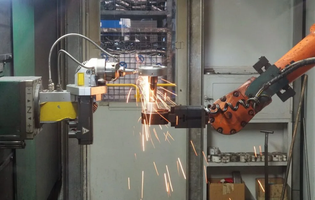 Un braccio robotico industriale arancione esegue una saldatura a scintille in un ambiente manifatturiero.