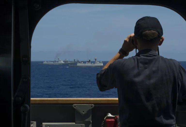 Militare in divisa scura osserva dal ponte di una nave una grande petroliera grigia in mare aperto.