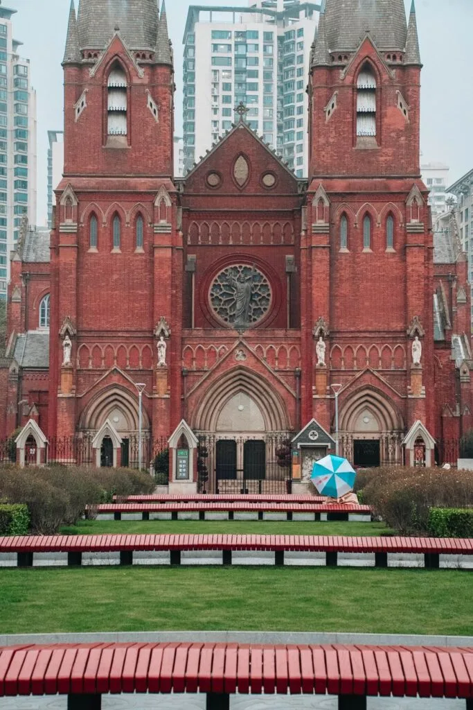 Cattedrale gotica in mattoni rossi con torri gemelle, circondata da grattacieli moderni e con ombrello blu in primo piano.