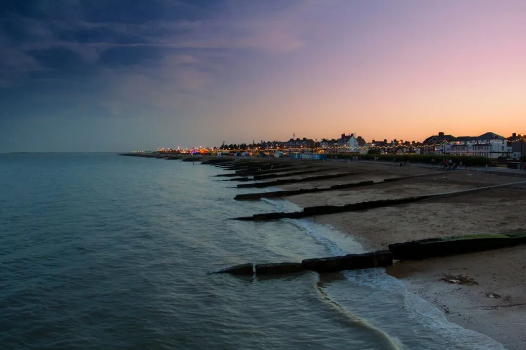 Spiaggia al tramonto con frangiflutti in legno e case illuminate sullo sfondo in una tranquilla costa inglese.