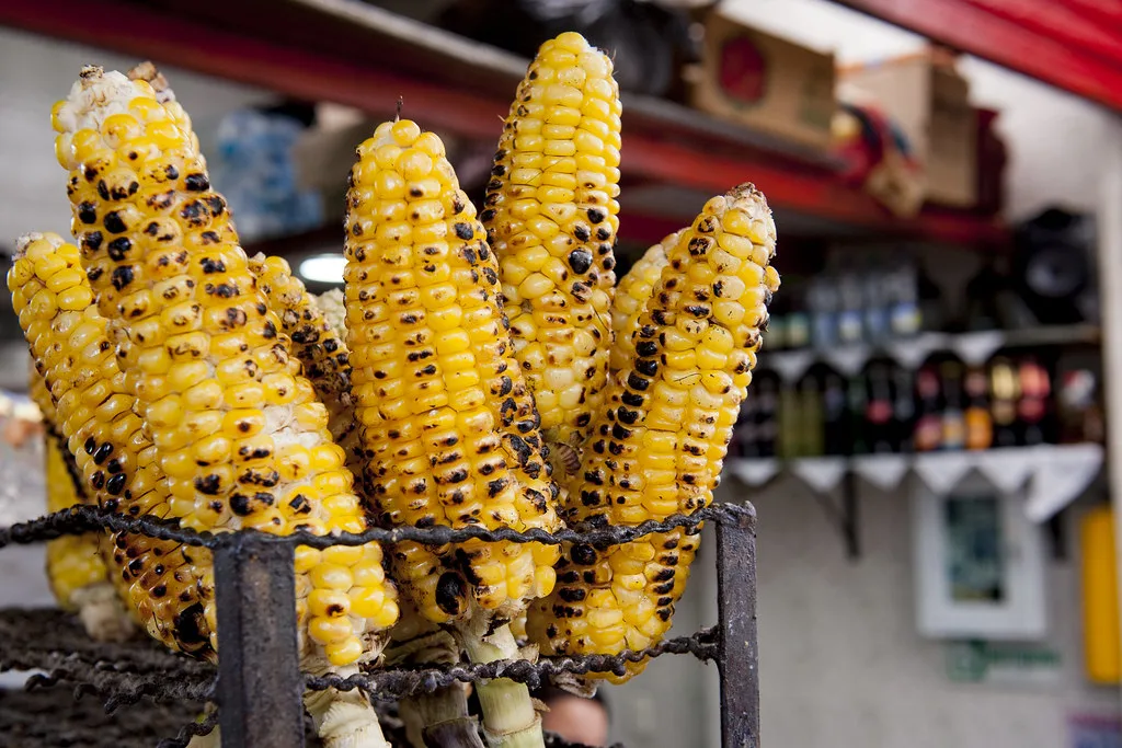 Pannocchie di mais giallo con tracce di carbonizzazione, esposte su un mercato locale con sfondo sfocato di bancarelle.