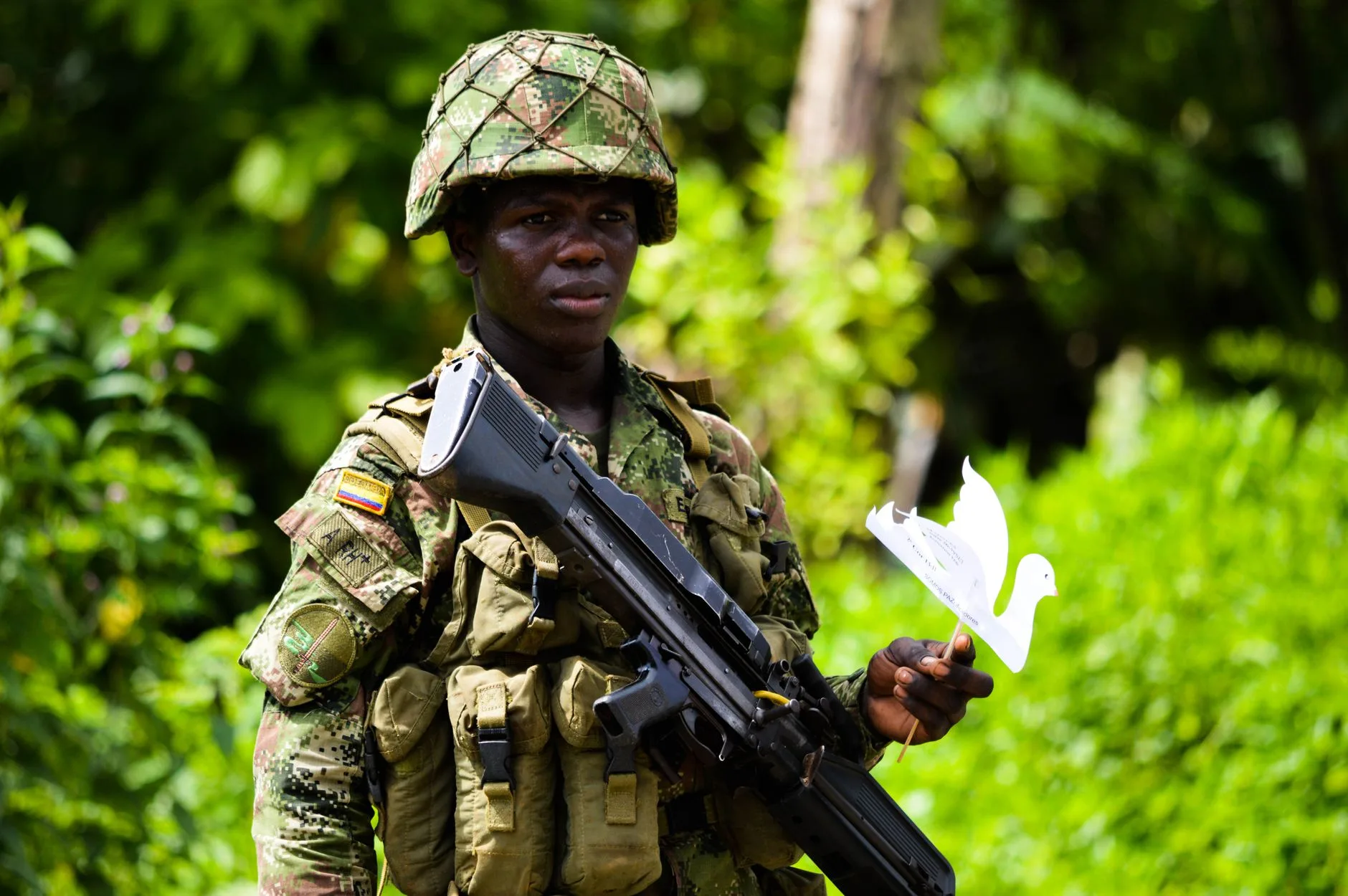 Soldato colombiano in uniforme mimetica con fucile e colomba di carta tra la vegetazione della giungla.