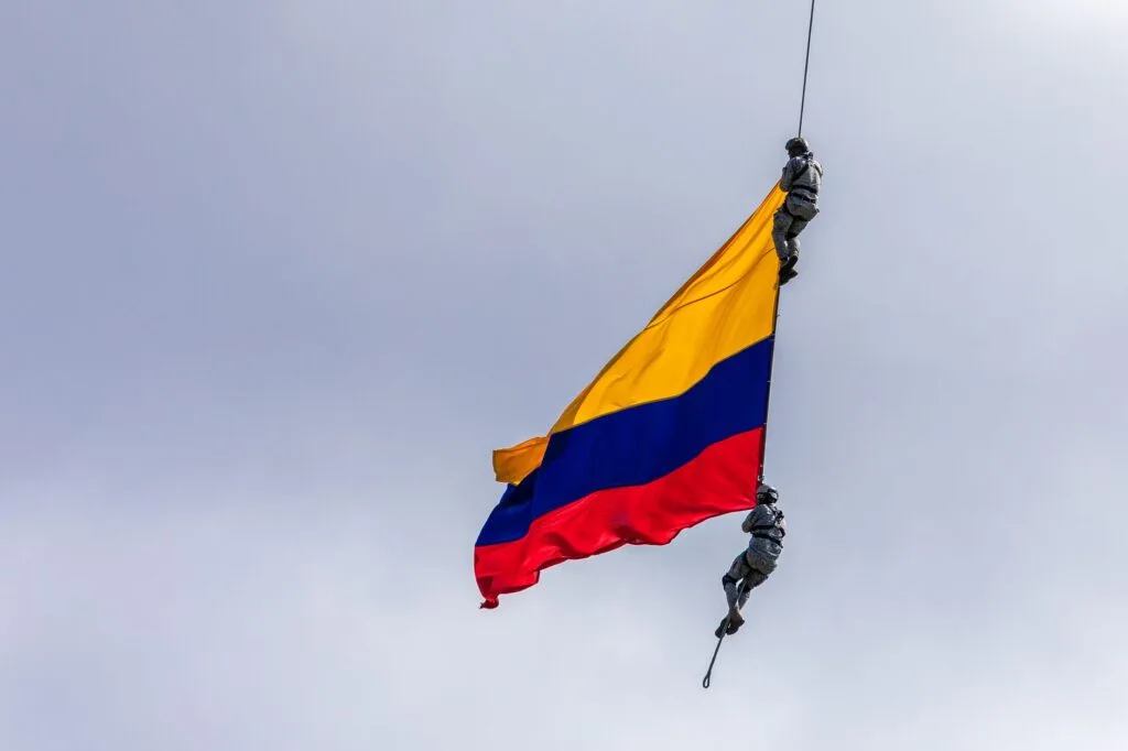 Due militari in calo da corda reggono la bandiera colombiana gialla-blu-rossa contro un cielo nuvoloso.
