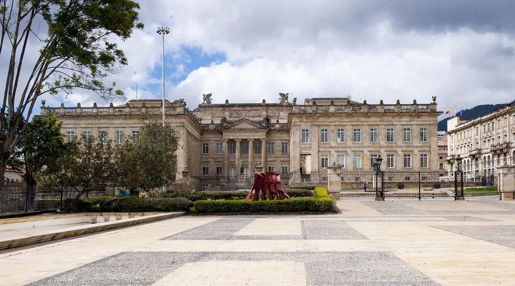 Palazzo neoclassico con colonne in piazza urbana circondata da edifici storici e vegetazione.