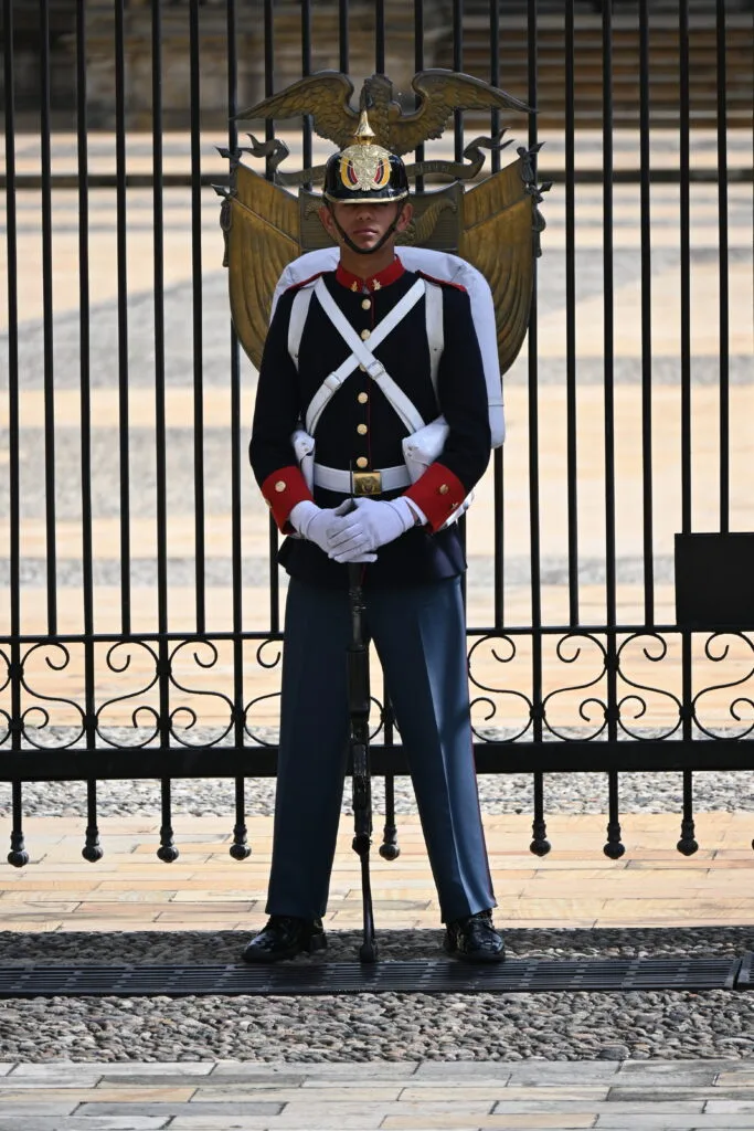 Guardia in uniforme cerimoniale colombiana con elmo piumato, davanti a cancellata ornamentale.