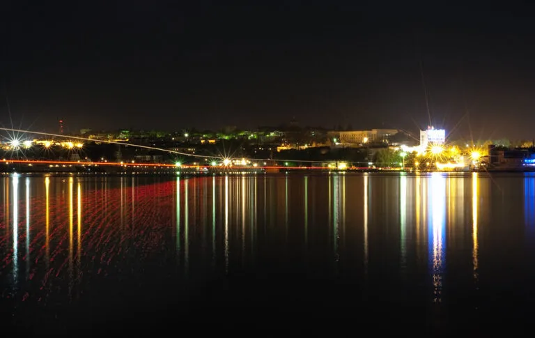 Città costiera illuminata di notte riflessa sull'acqua scura, con luci gialle e bianche che si specchiano nel mare.