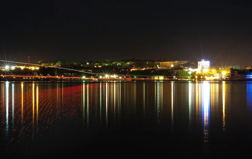 Città costiera illuminata di notte riflessa sull'acqua scura, con luci gialle e bianche che si specchiano nel mare.