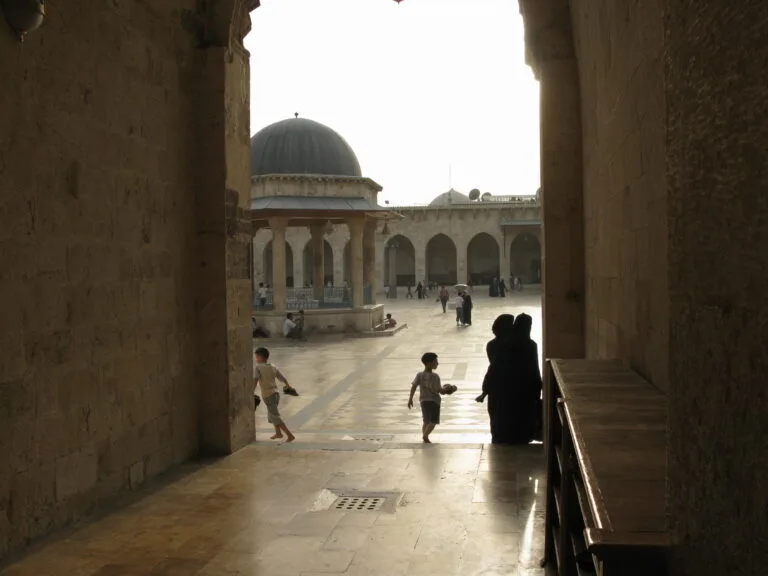 Cortile di una grande moschea con arcate e cupola, alcune persone camminano sul pavimento bagnato.