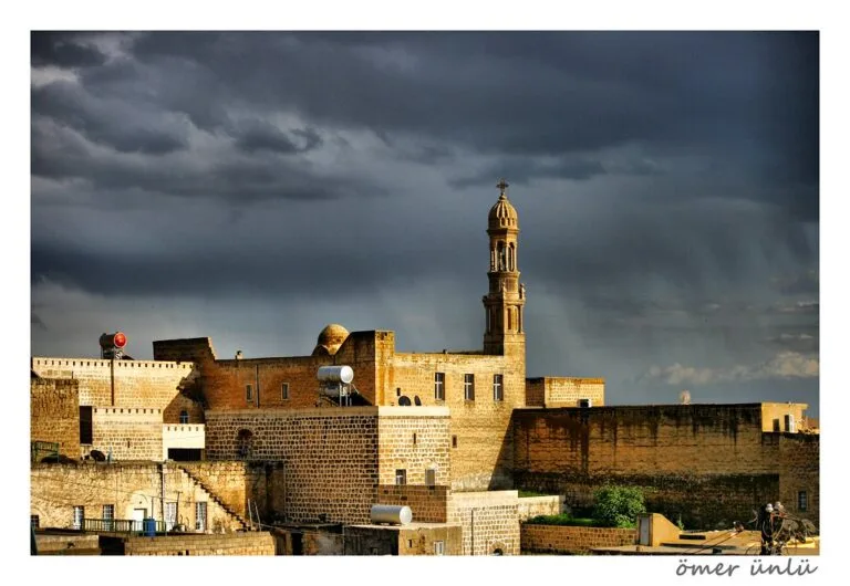 Fortificazione medievale con torre merlata illuminata dal sole al tramonto, sotto cielo nuvoloso scuro.