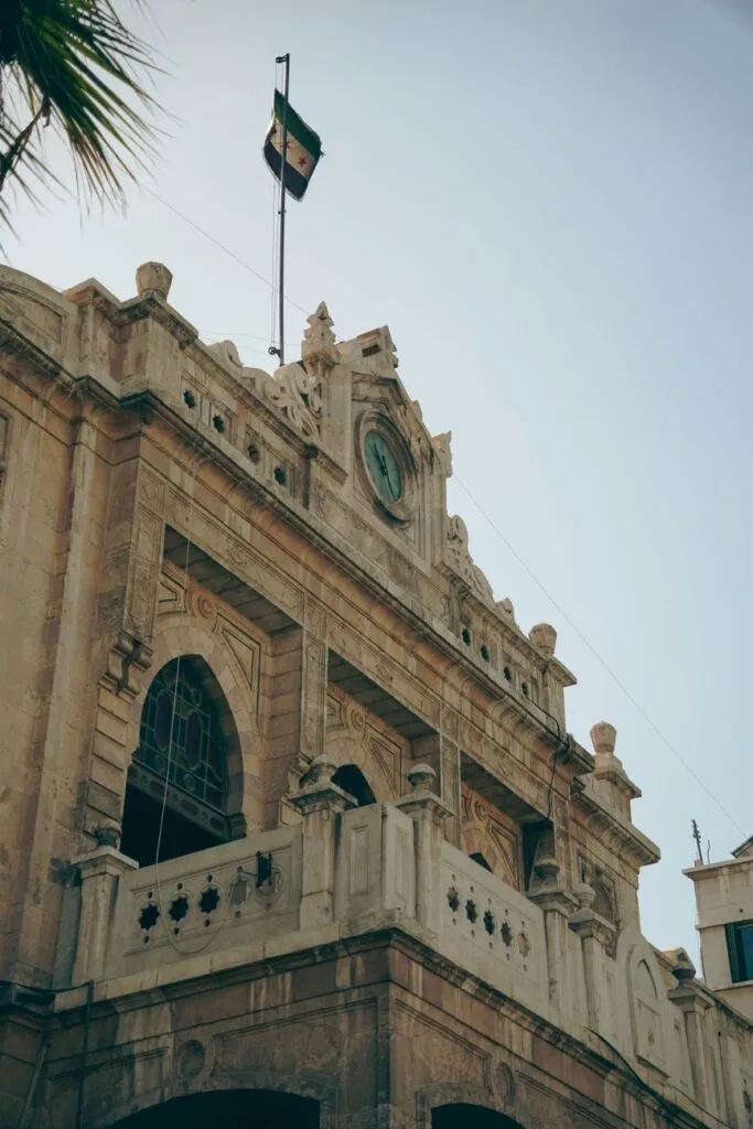 Edificio storico in pietra con orologio, balconate decorate e bandiera nazionale siriana issata in cima.
