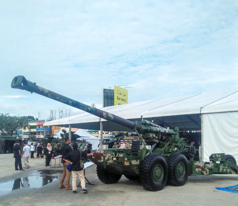Cannone semovente verde su piattaforma a sei ruote esposto in una fiera con visitatori e strutture commerciali sullo sfondo.