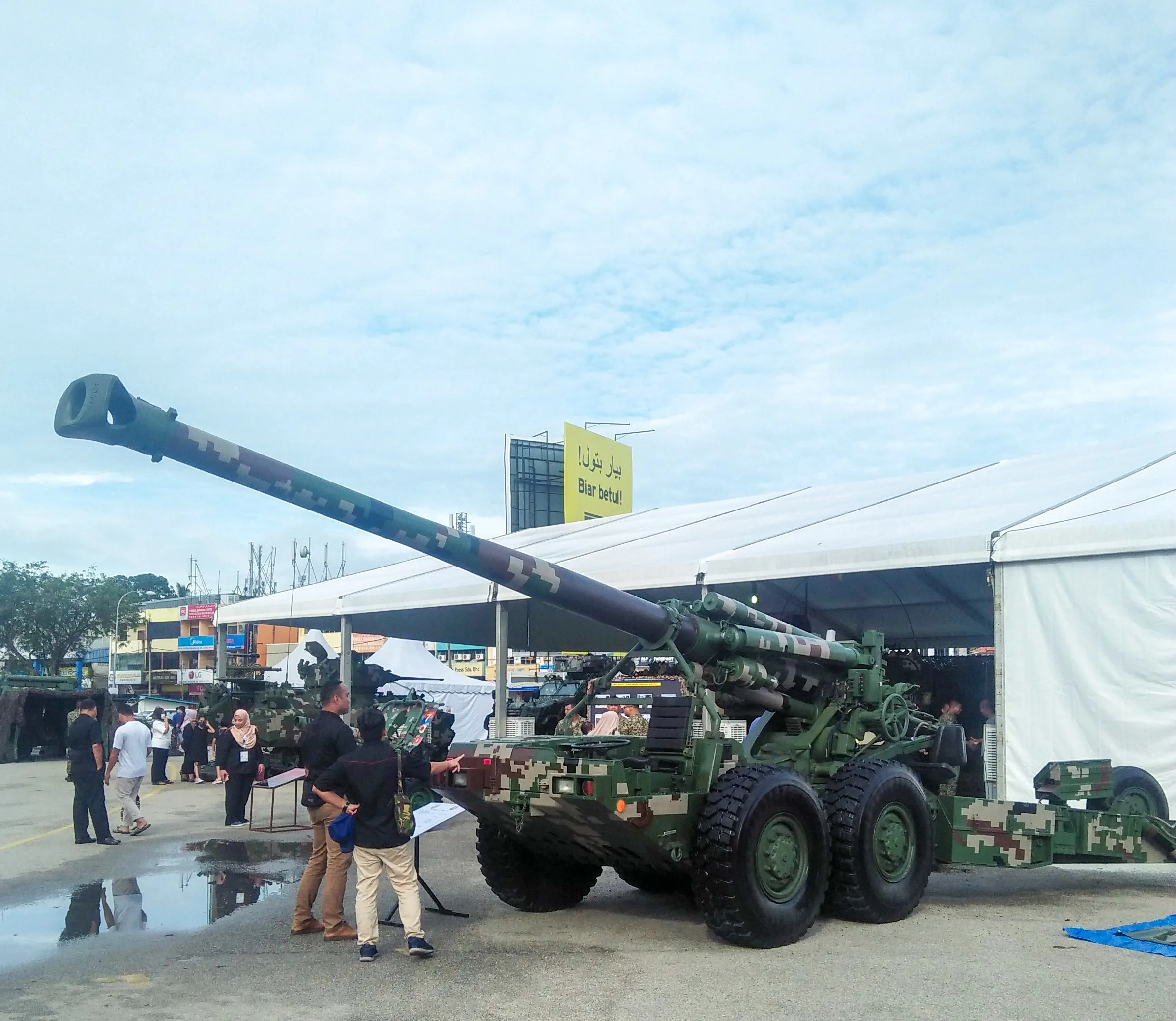 Cannone semovente verde su piattaforma a sei ruote esposto in una fiera con visitatori e strutture commerciali sullo sfondo.