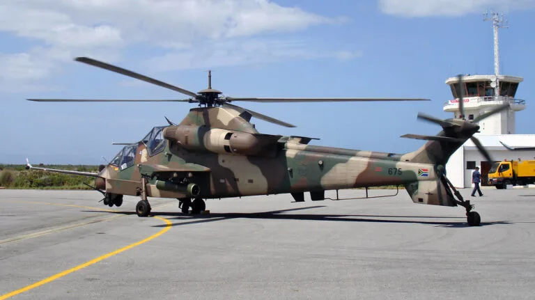 Elicottero da attacco Rooivalk con mimetismo verde-marrone parcheggiato in aeroporto militare accanto a torre di controllo.