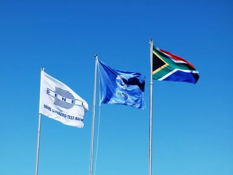 Tre bandiere sventolano su pali contro cielo blu: quella del Sudafrica, una bandiera blu e una bandiera istituzionale.