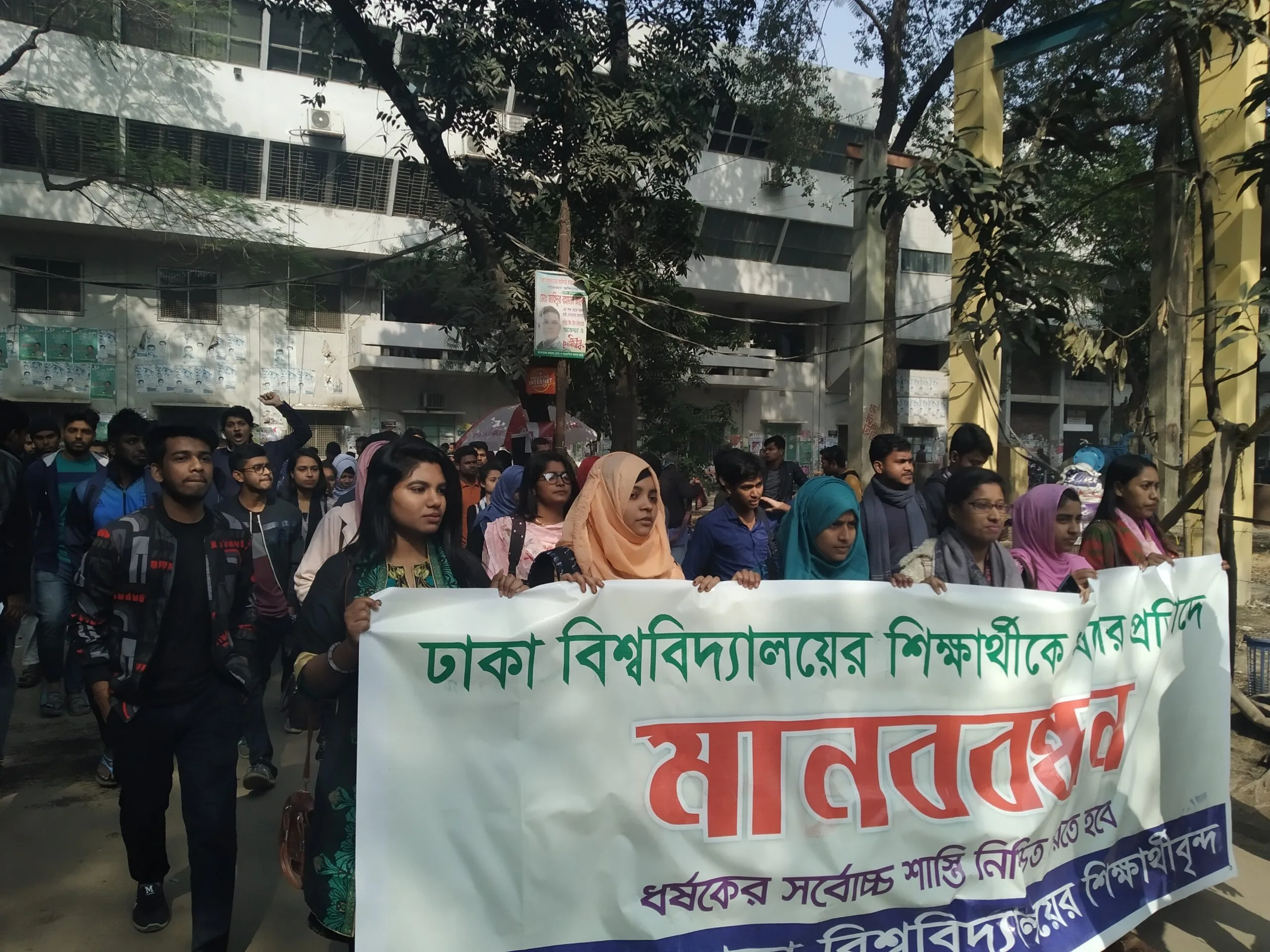 Studenti bangladesi manifestano in strada con striscioni in bengali davanti a edifici universitari.