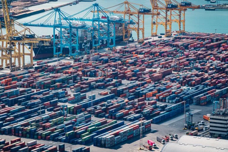 Porto marittimo con migliaia di container rossi e blu impilati, gru di carico e nave CGA attraccata al molo.