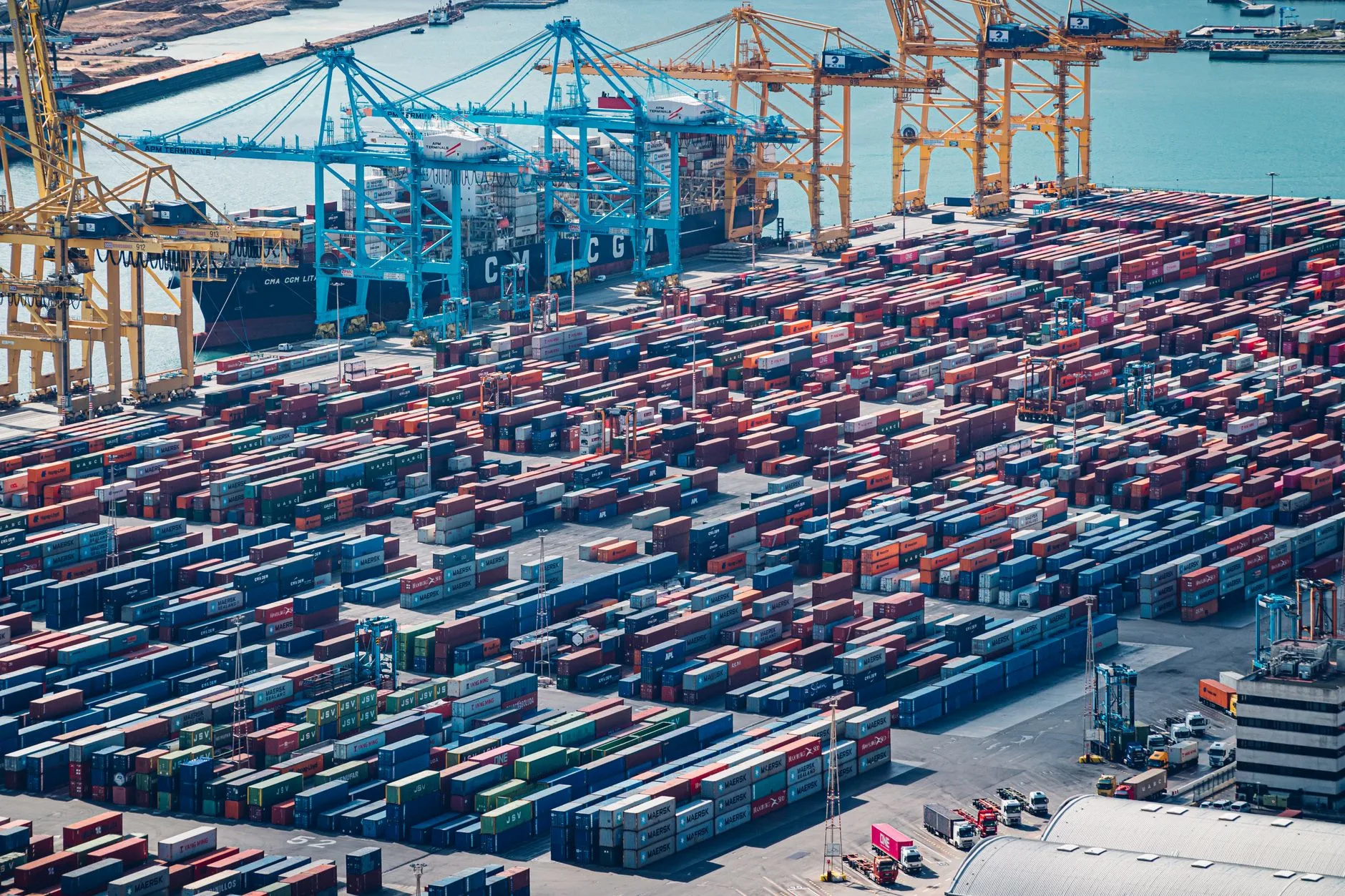 Porto marittimo con migliaia di container rossi e blu impilati, gru di carico e nave CGA attraccata al molo.
