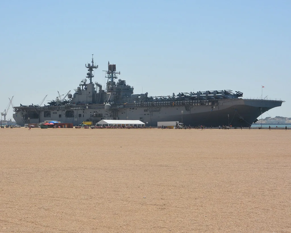 Portaelicotteri militare attraccata in porto con container e attrezzature di servizio lungo la costa sabbiosa.